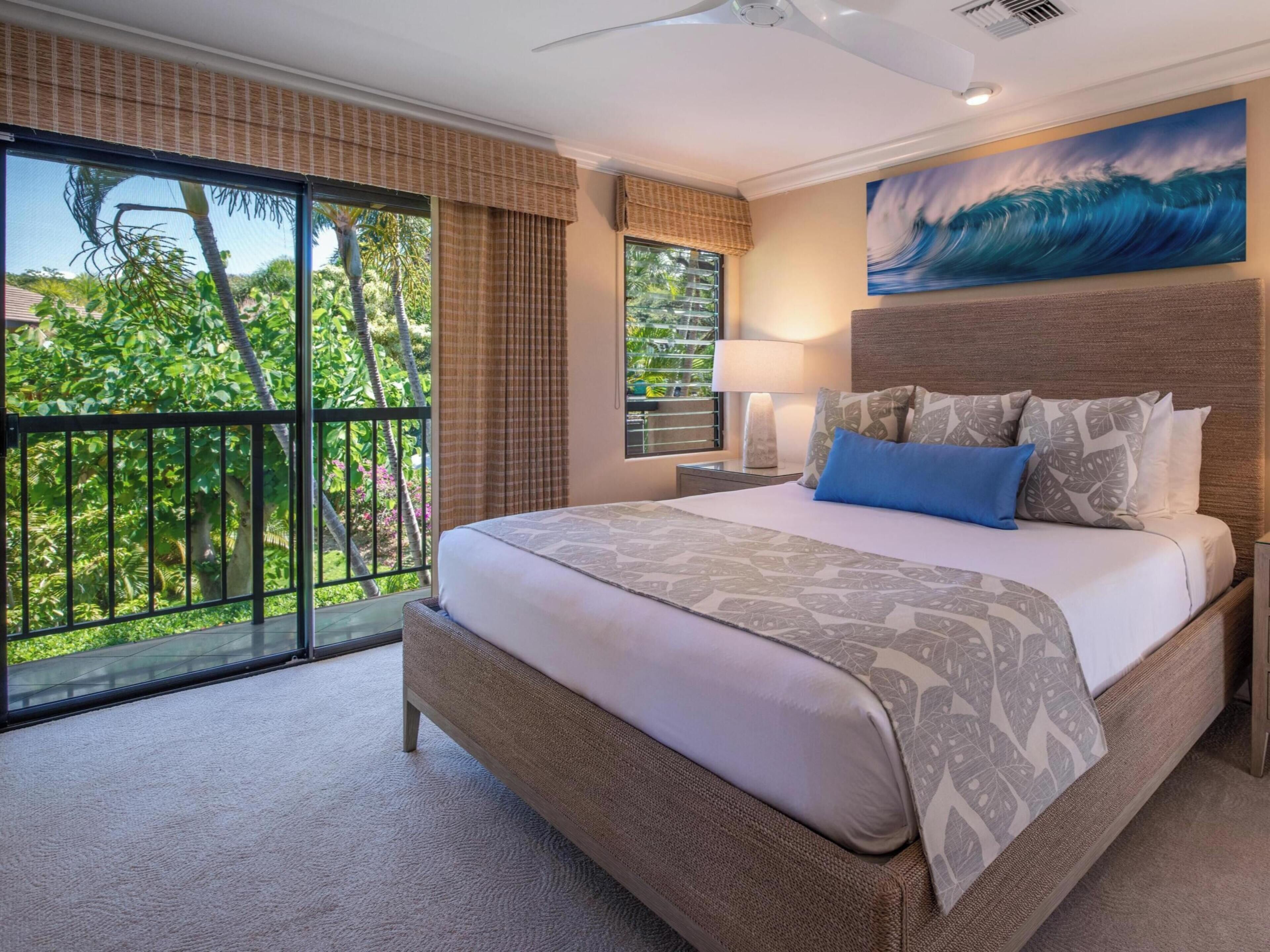A spacious bedroom with a large bed, a view of lush greenery through floor-to-ceiling windows, and a painting of a wave on the wall.