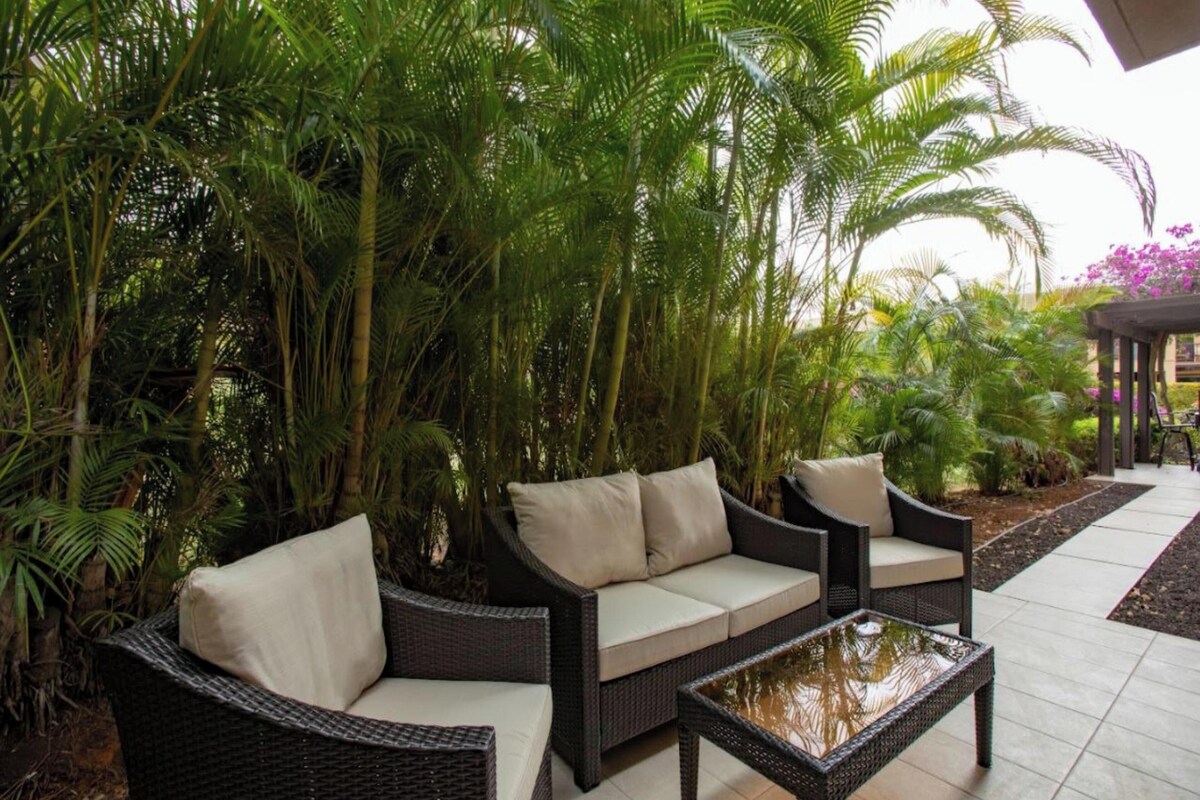A patio area with wicker furniture and a glass-top coffee table, surrounded by lush tropical plants.