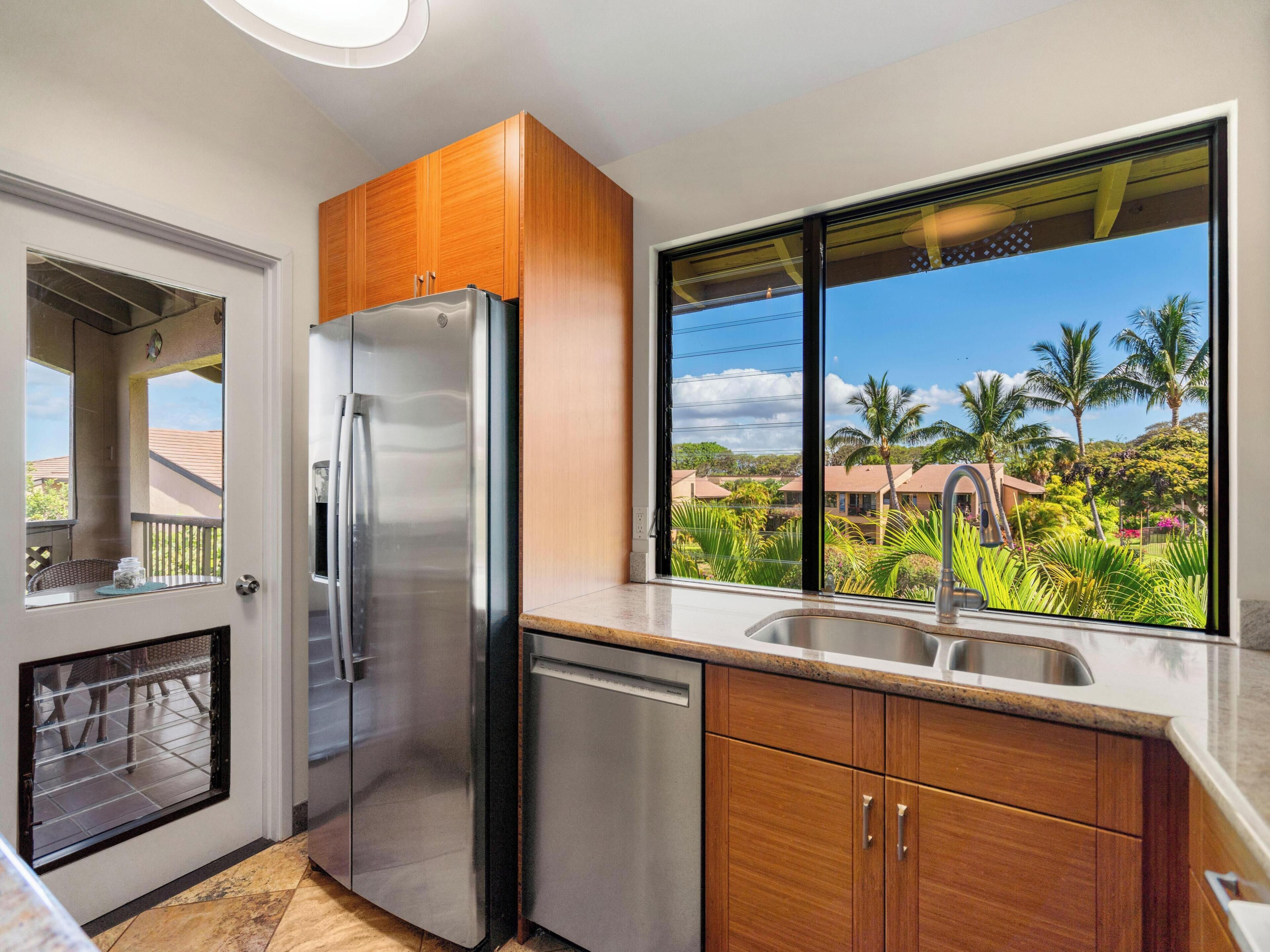 A kitchen with a stainless steel refrigerator, wooden cabinets, and a large window offering a view of a residential area with palm trees.