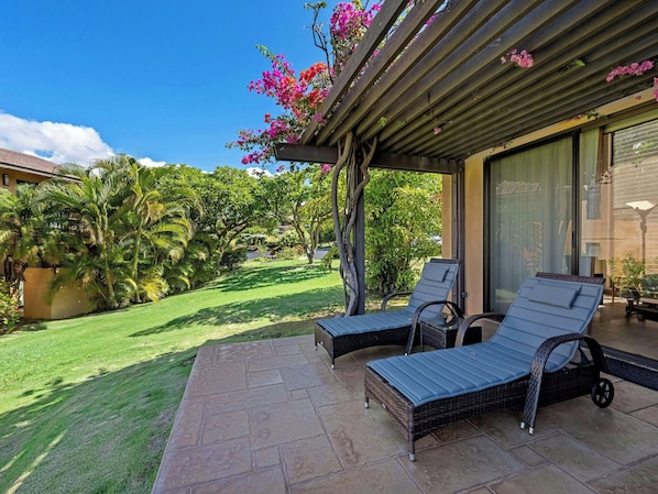 A patio with two lounge chairs, a pergola with flowering vines, and a view of a lush garden.