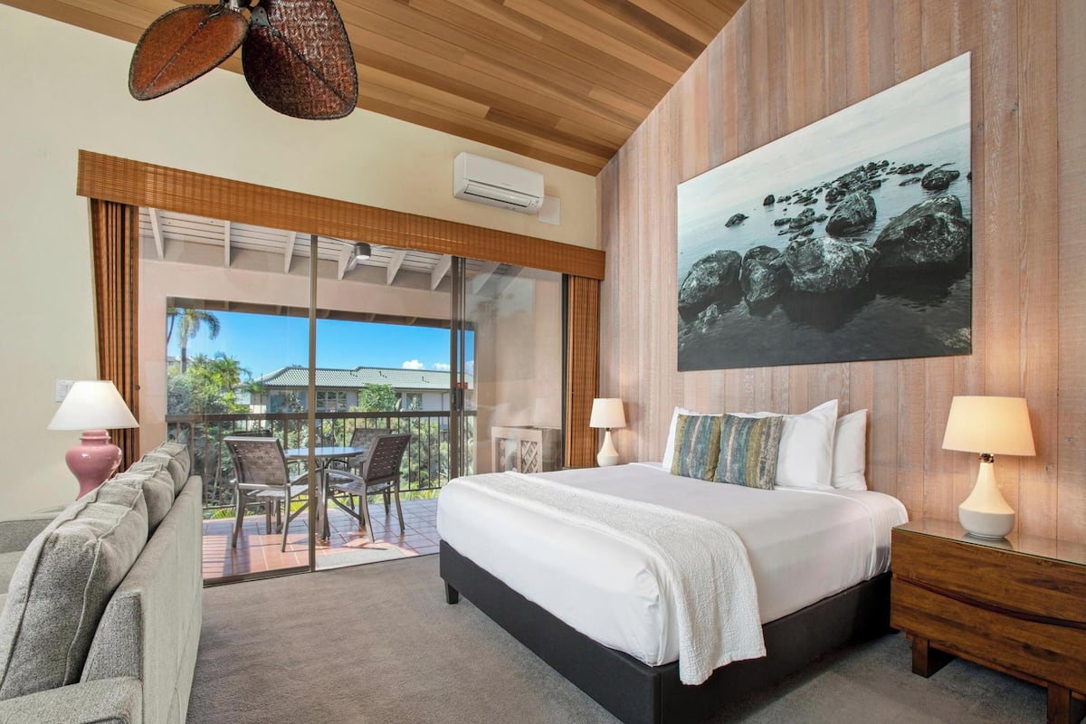 Un cuarto de hotel con una cama grande, un balcón con vista a palmeras y un cuadro de rocas en la pared.
