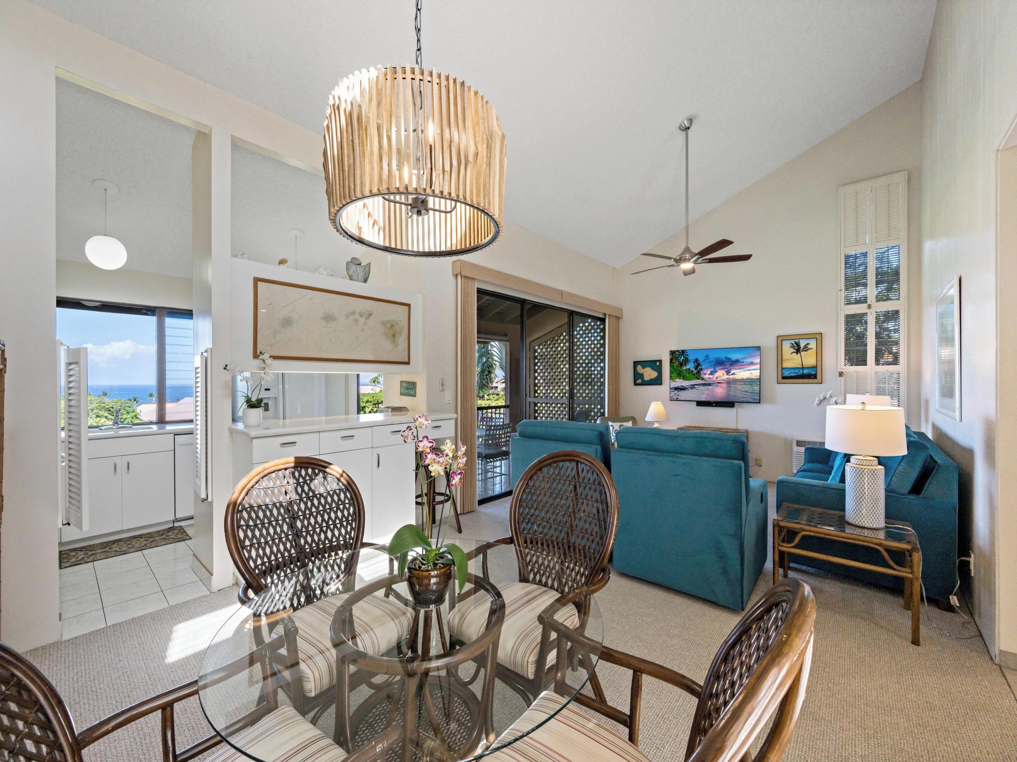 A spacious living area with a dining table, chairs, and a large blue sofa. There is a ceiling fan, a TV mounted on the wall, and a view of the ocean through a window.