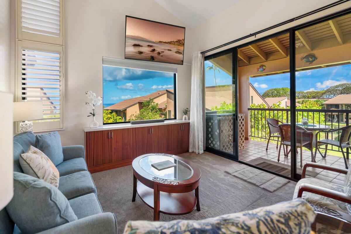 Una sala de estar con sofá, una mesa de centro y un ventanal con vista a una playa y casas.
