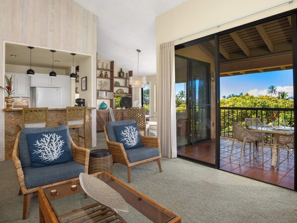 A spacious living area with wicker furniture, a glass coffee table, and a view of a balcony with outdoor seating.