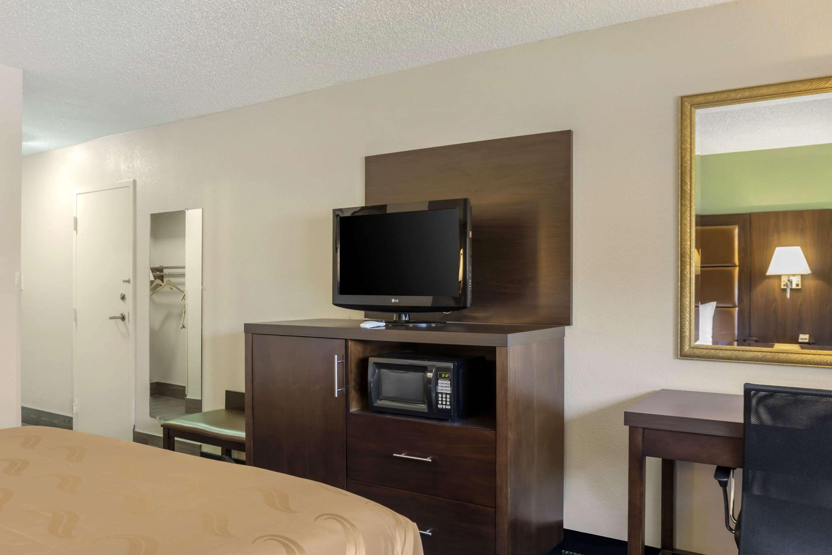 A hotel room with a bed, a TV on a wooden cabinet, a microwave, a desk with a chair, and a mirror on the wall.