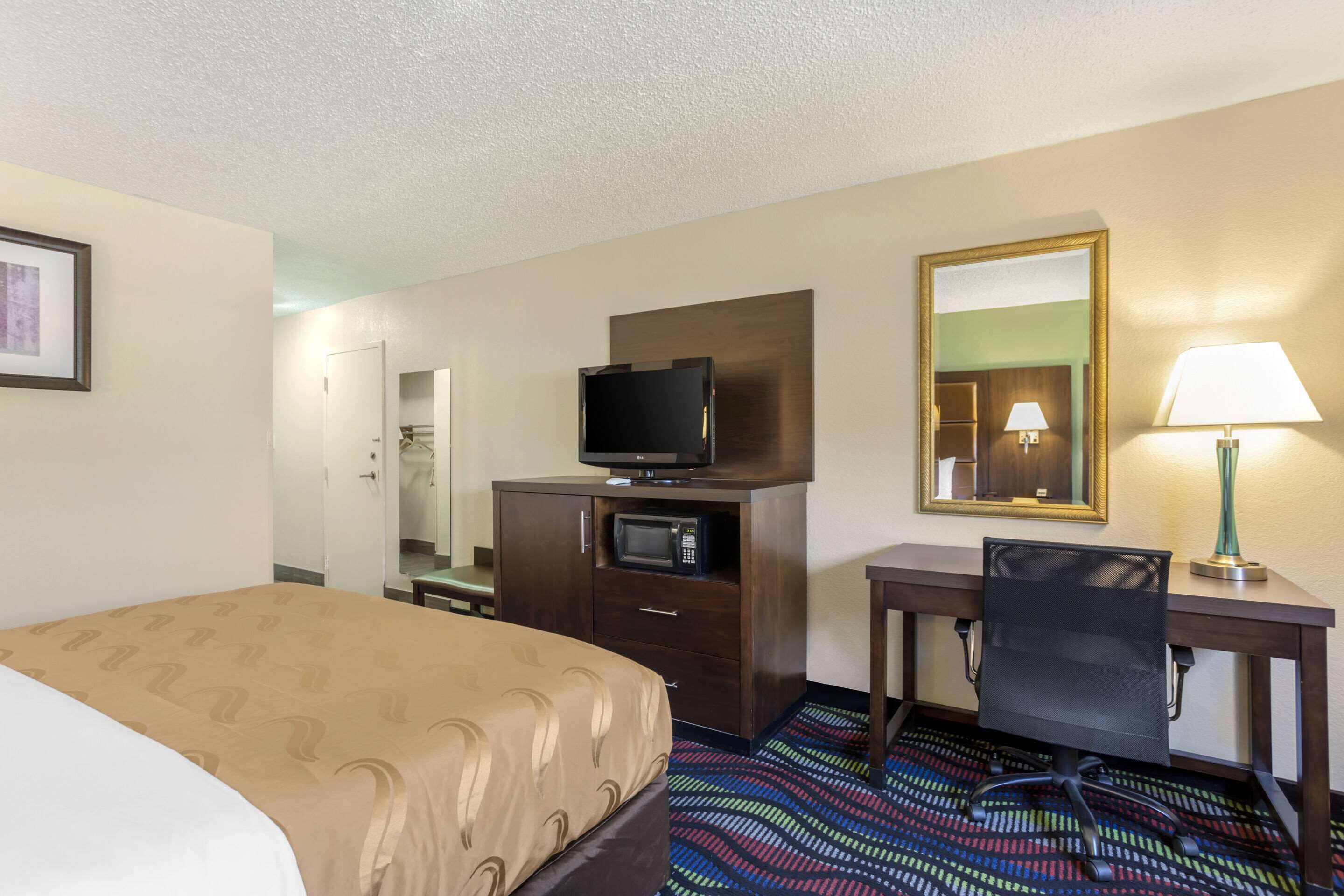 A hotel room with a bed, a desk with a chair, a television, a microwave, a lamp, and a mirror.