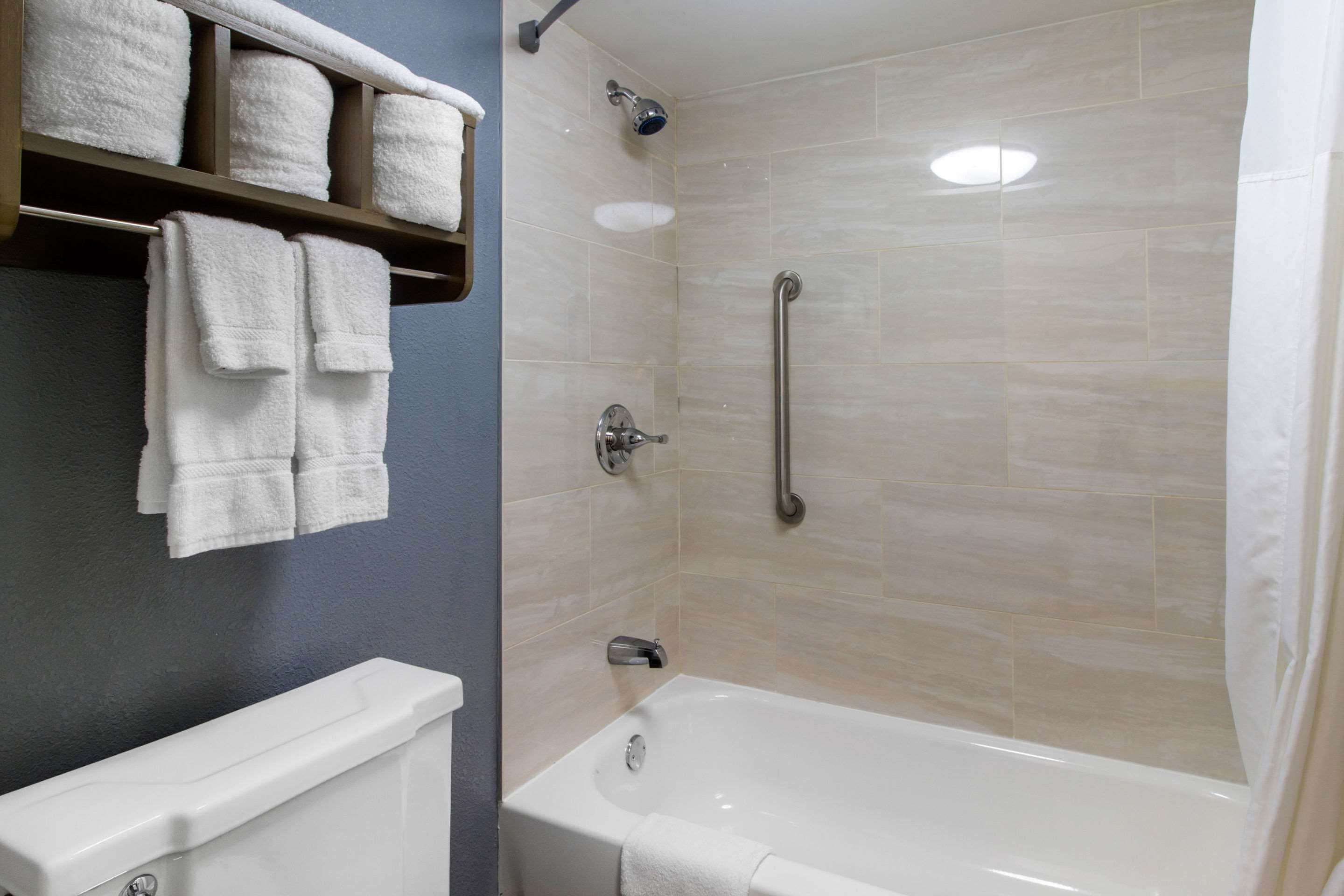 A bathroom with a bathtub, a toilet, a shelf with towels, and a shower with a grab bar.