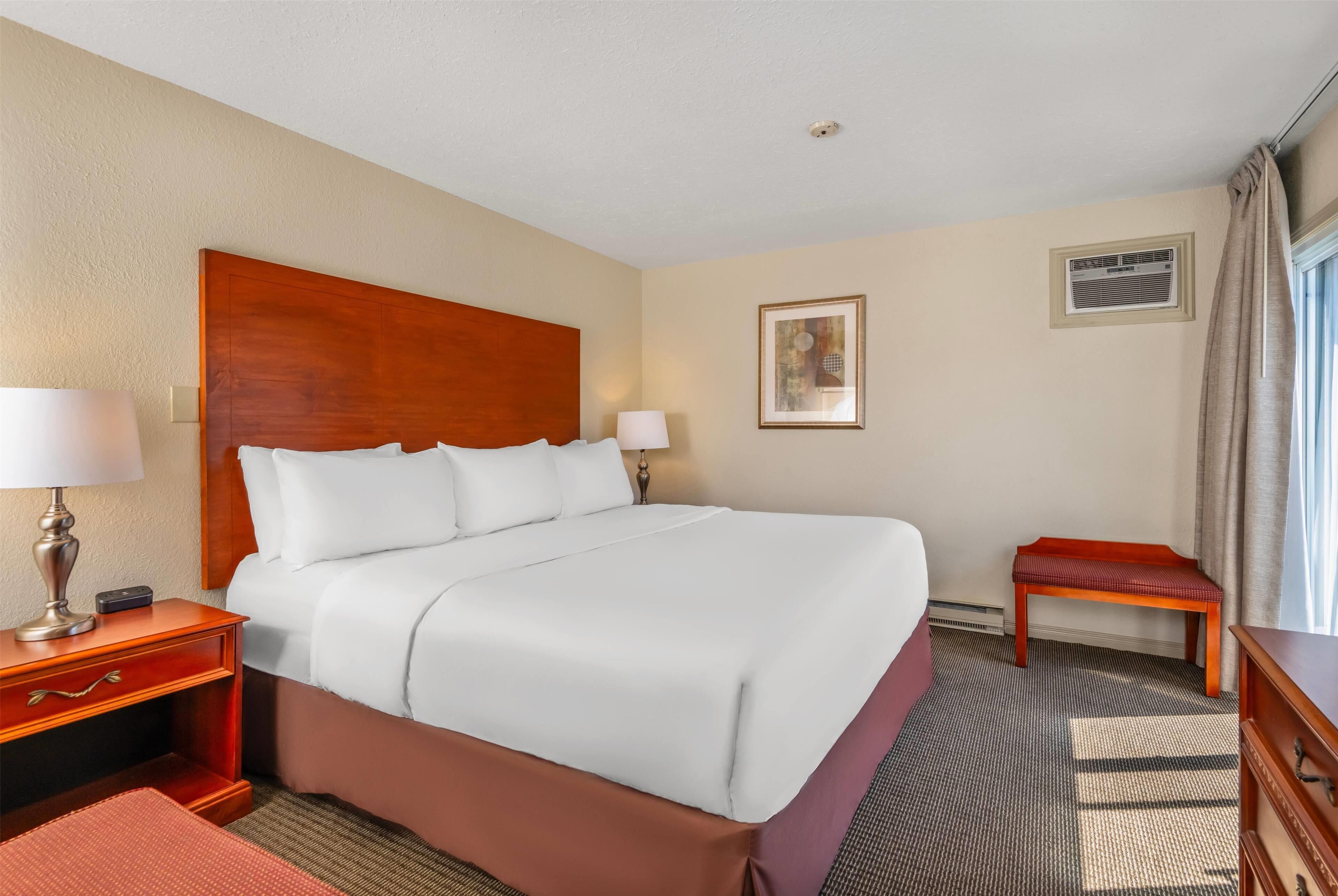 A hotel room with a large bed, a nightstand with a lamp, a small table, a bench, and a framed picture on the wall.