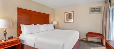A hotel room with a large bed, a nightstand with a lamp, a small table, a bench, and a framed picture on the wall.