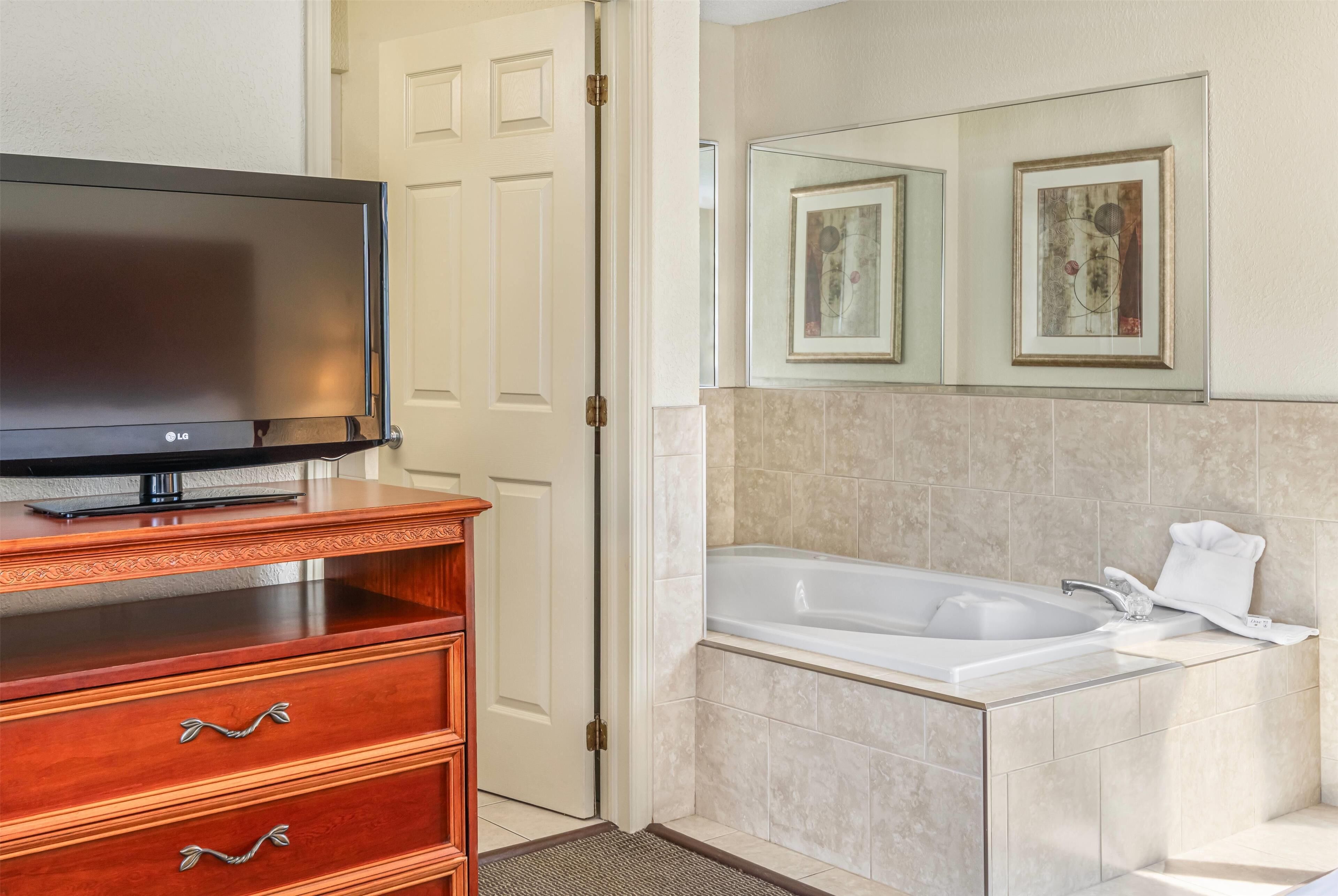 A bathroom with a bathtub, a television on a wooden dresser, and framed artwork on the wall.
