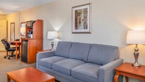 Ein Hotelzimmer mit grauem Sofa, einem Holztischchen, einem Schreibtisch mit Computer, einem Sessel und einem gerahmten Bild an der Wand.