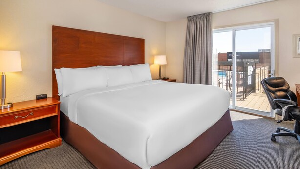 Ein Hotelzimmer mit einem großen Bett, einem Schreibtisch mit Lampe, einem Sessel und Blick auf ein Gebäude sowie einen Pool durch eine Glastür.