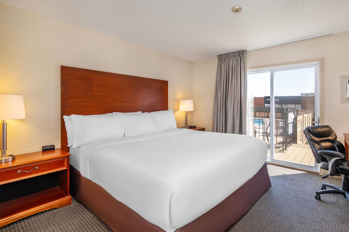 Une chambre d’hôtel avec un grand lit, un bureau avec une lampe, une chaise et une vue sur un immeuble et une piscine, visibles à travers une porte vitrée.