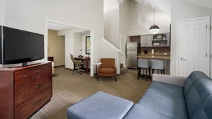 A modern living room with a flat-screen TV, a sofa, a coffee table, and a dining area with a kitchen in the background.