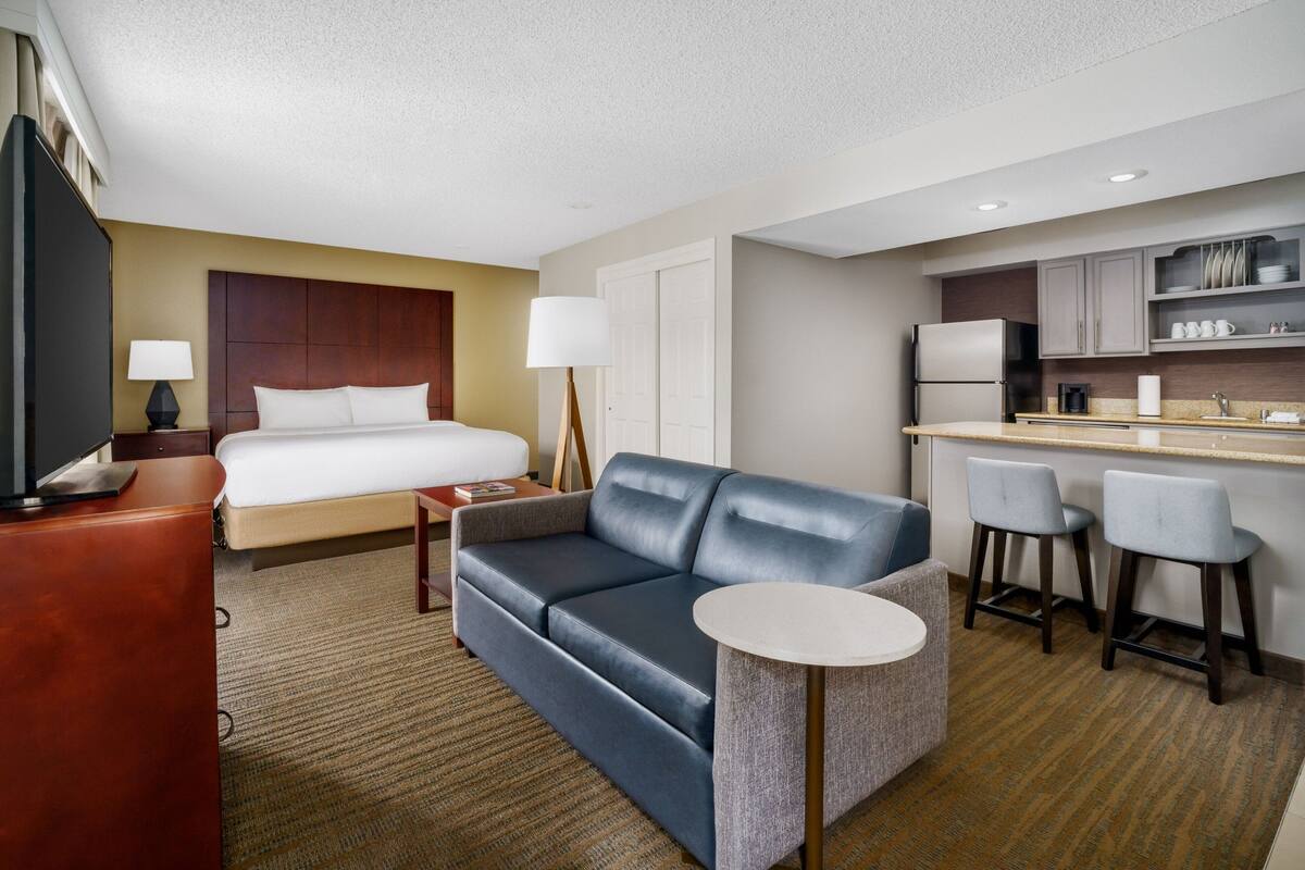A hotel room with a bed, a sofa, a small table, a TV, a desk, and a kitchenette.