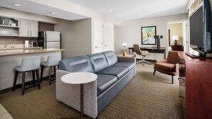 A modern hotel room with a kitchenette, a grey sofa, a small round table, a TV, and a desk with a chair.