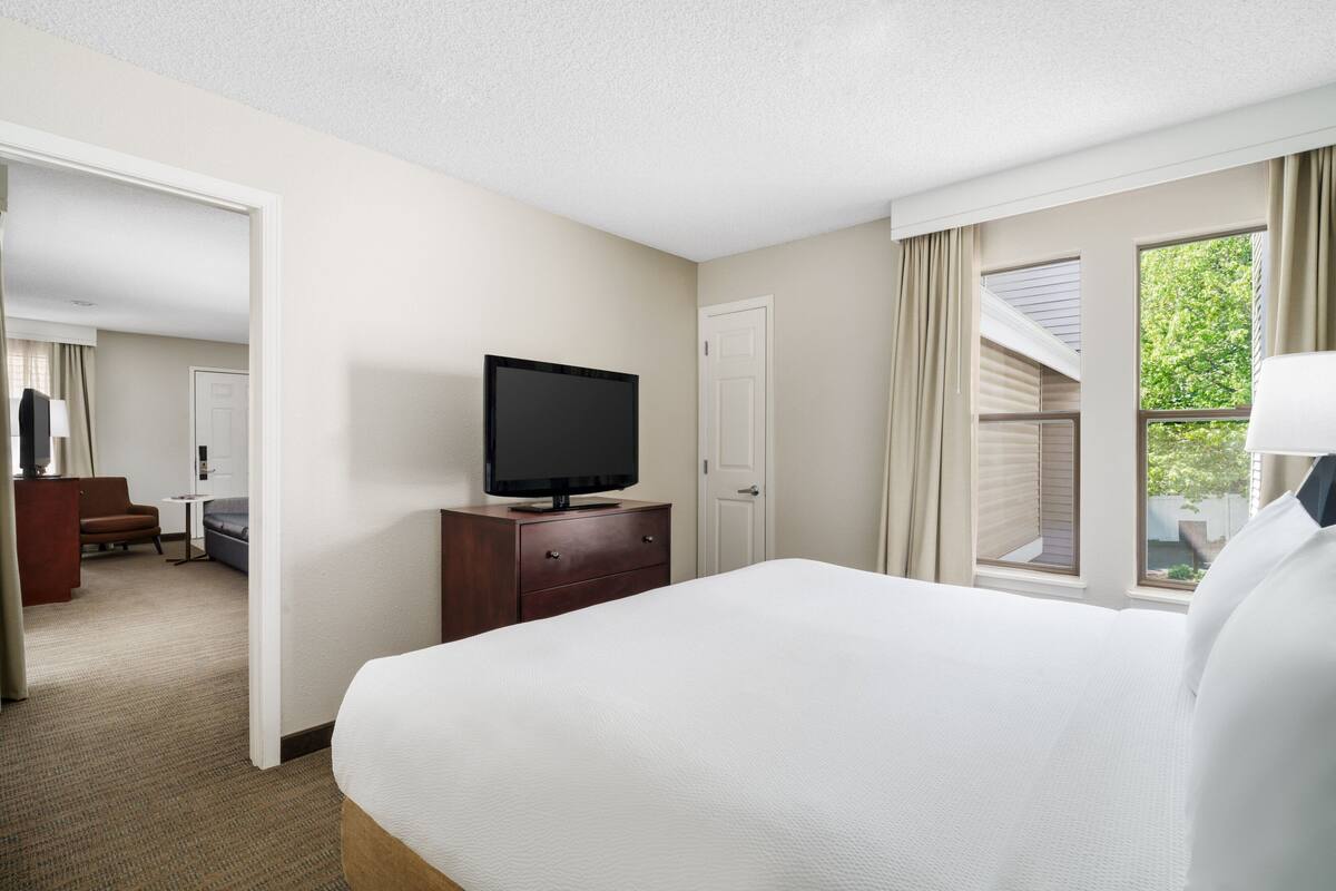 A hotel room with a large bed, a television on a dresser, a desk, and a window with curtains.