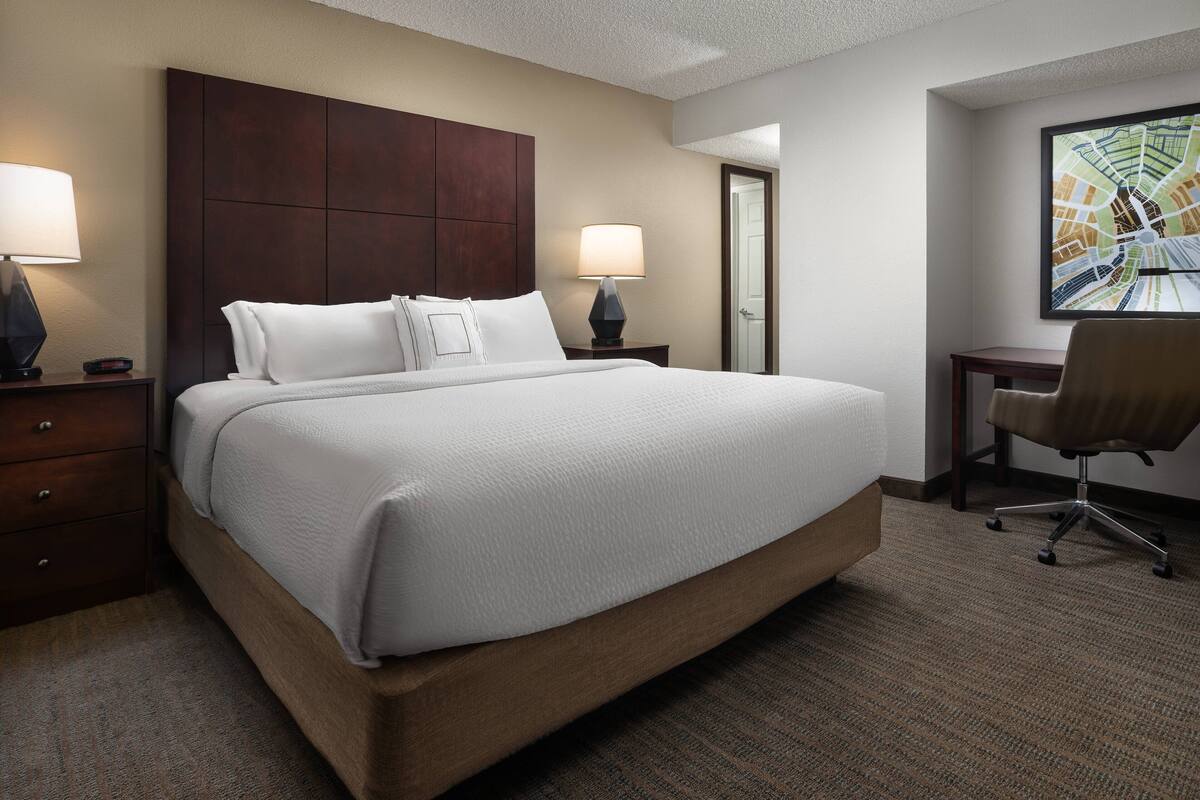 A hotel room with a large bed, two bedside tables with lamps, a desk with a chair, and a framed cityscape artwork on the wall.