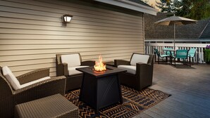 An outdoor patio with wicker furniture, a square table with a fire feature, and a patterned rug.