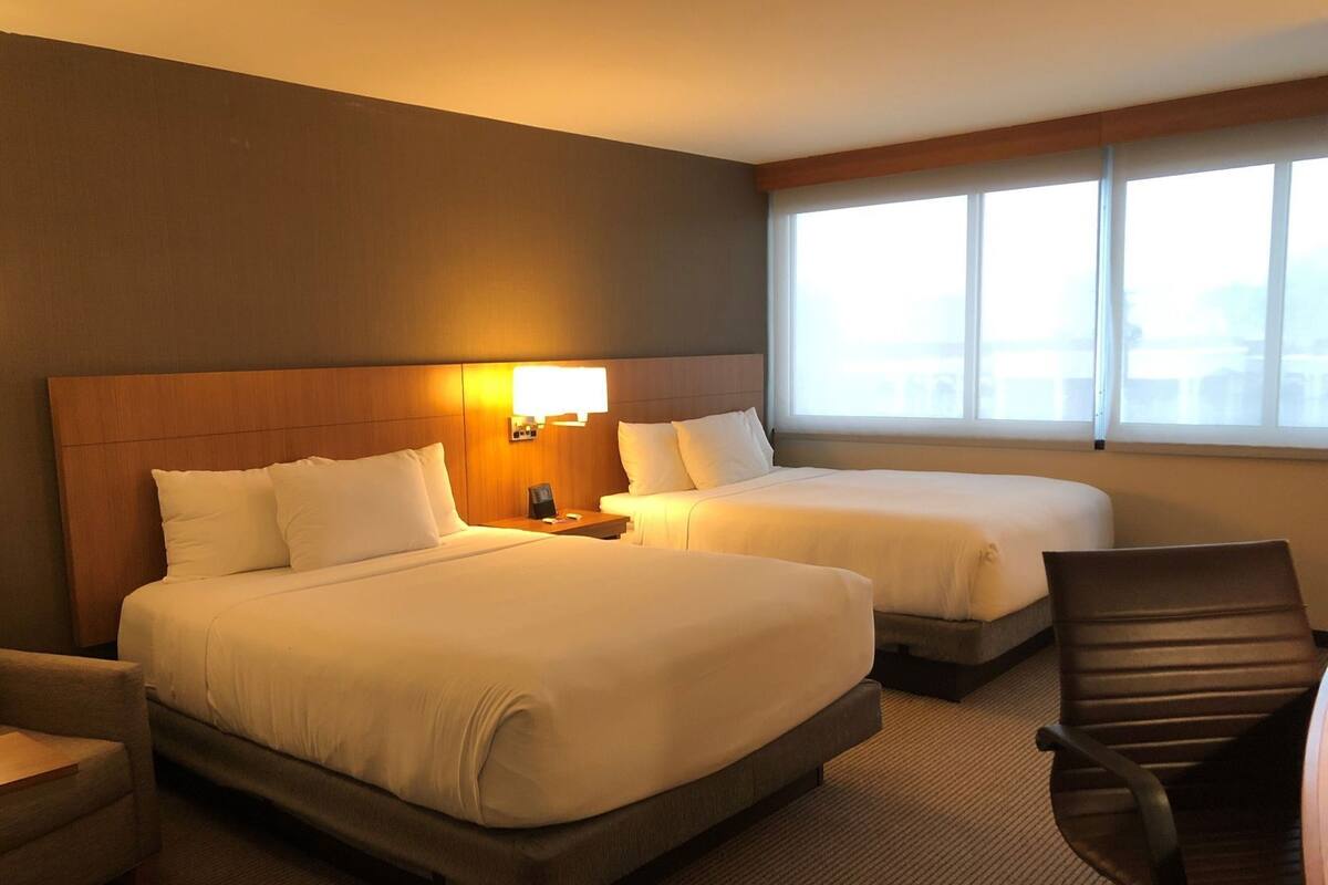 A hotel room with two beds, a nightstand, a chair, and a large window.