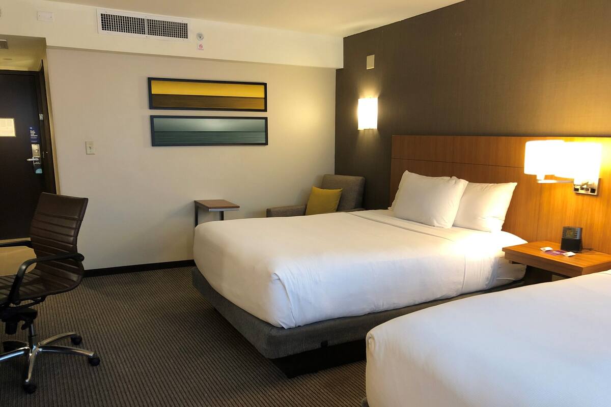 A hotel room with two beds, a desk, a chair, and a wall-mounted light.