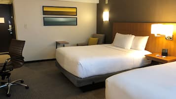 A hotel room with two beds, a desk, a chair, and a wall-mounted light.