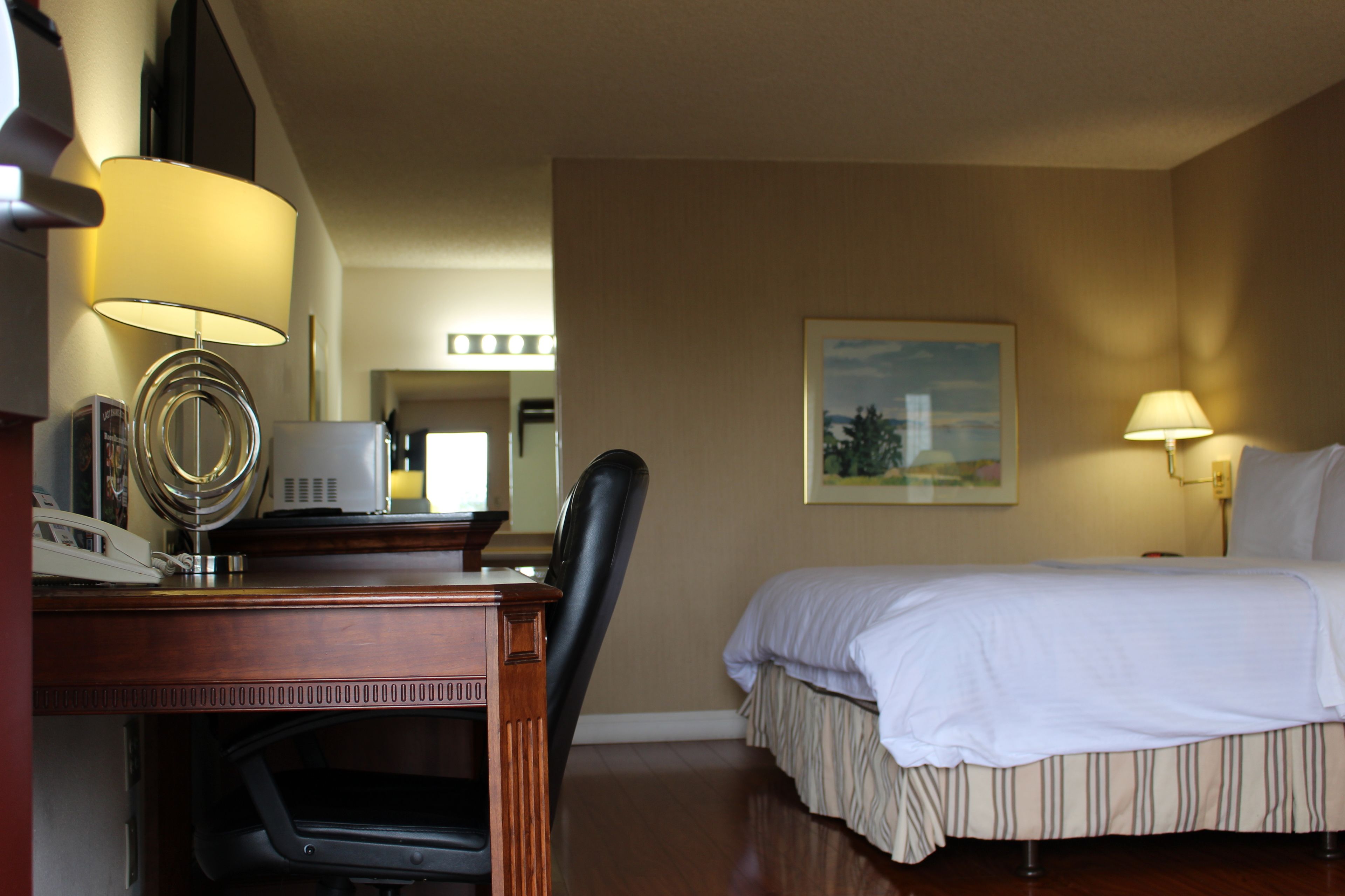 A hotel room with a bed, desk, chair, lamp, and a painting on the wall.