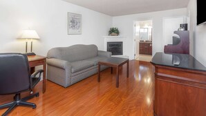 A living room with a gray sofa, wooden coffee table, and a desk with a lamp. There is a fireplace with a plant on top, and a TV mounted on the wall. - Days Inn & Suites by Wyndham Sunnyvale (Sunnyvale)