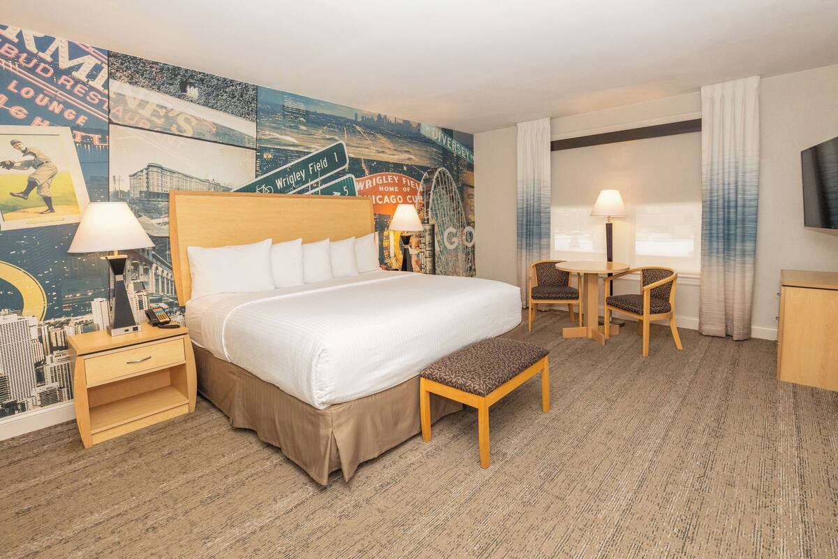 Une chambre d’hôtel avec un grand lit, des tables de chevet avec des lampes, un bureau avec une chaise, une télévision et une fresque représentant un paysage urbain sur le mur.