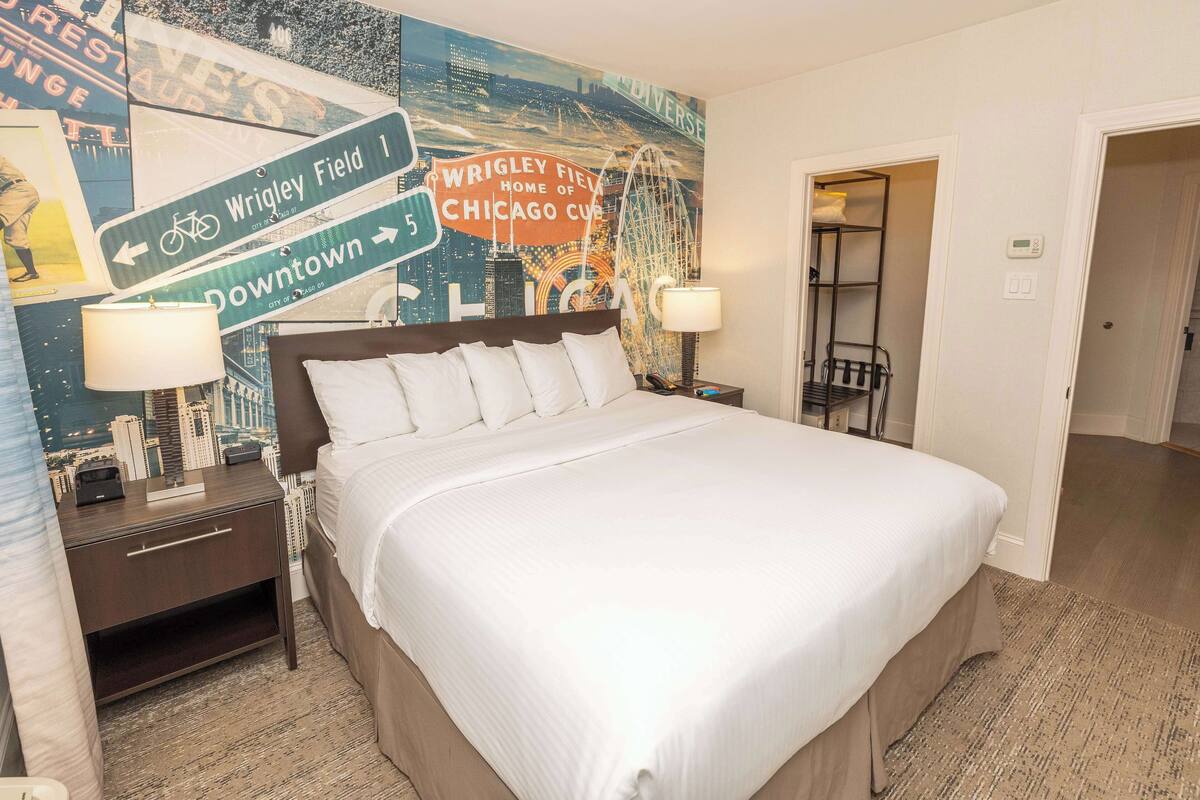 Une chambre d’hôtel avec un grand lit, une table de chevet avec une lampe, une fresque murale et une porte donnant sur une autre pièce.