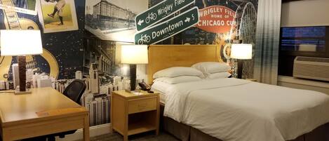 A hotel room with a bed, desk, and nightstand. There are posters on the wall, a lamp, and a window with curtains.