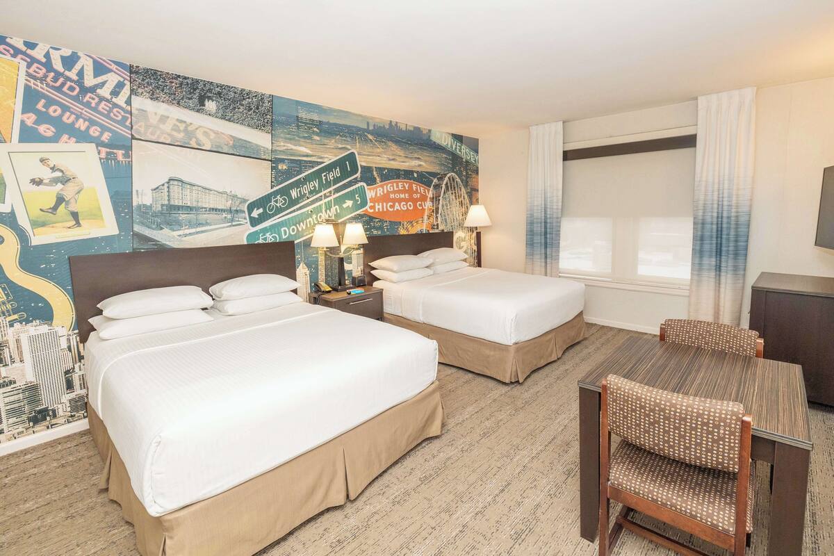 Une chambre d’hôtel avec deux lits, une table à manger et une chaise. Un mur est décoré d’une fresque représentant un paysage urbain et une grande roue.
