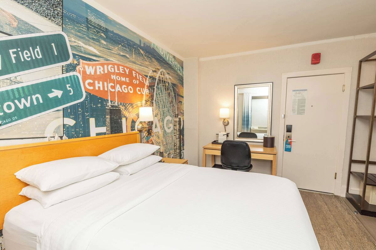 Une chambre d’hôtel avec un grand lit, un bureau avec une chaise, un miroir et un mur orné d’une fresque représentant un paysage urbain.