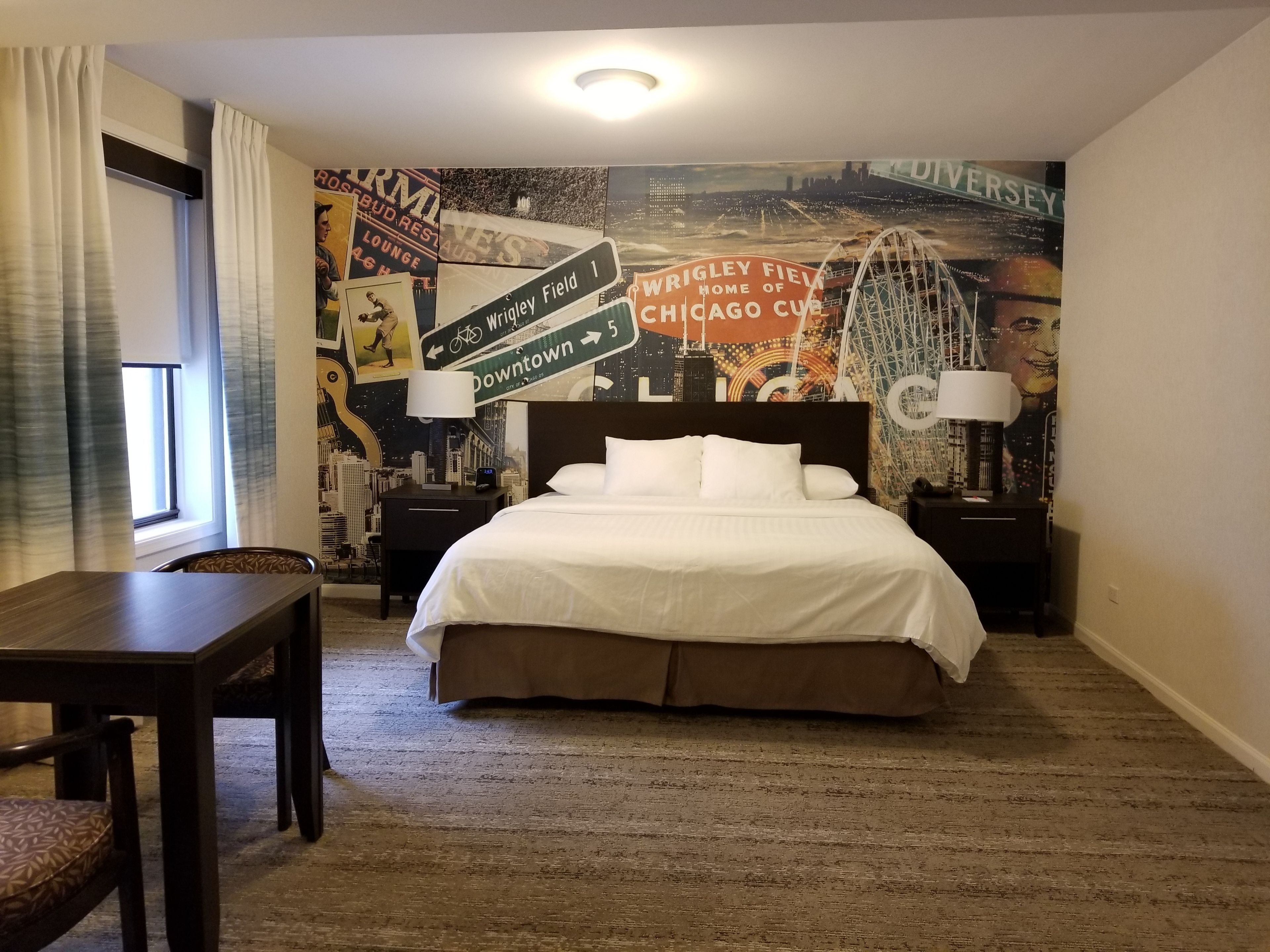 A hotel room with a large bed, two bedside tables with lamps, a desk, and a chair. The walls are decorated with vintage sports and cityscape posters.