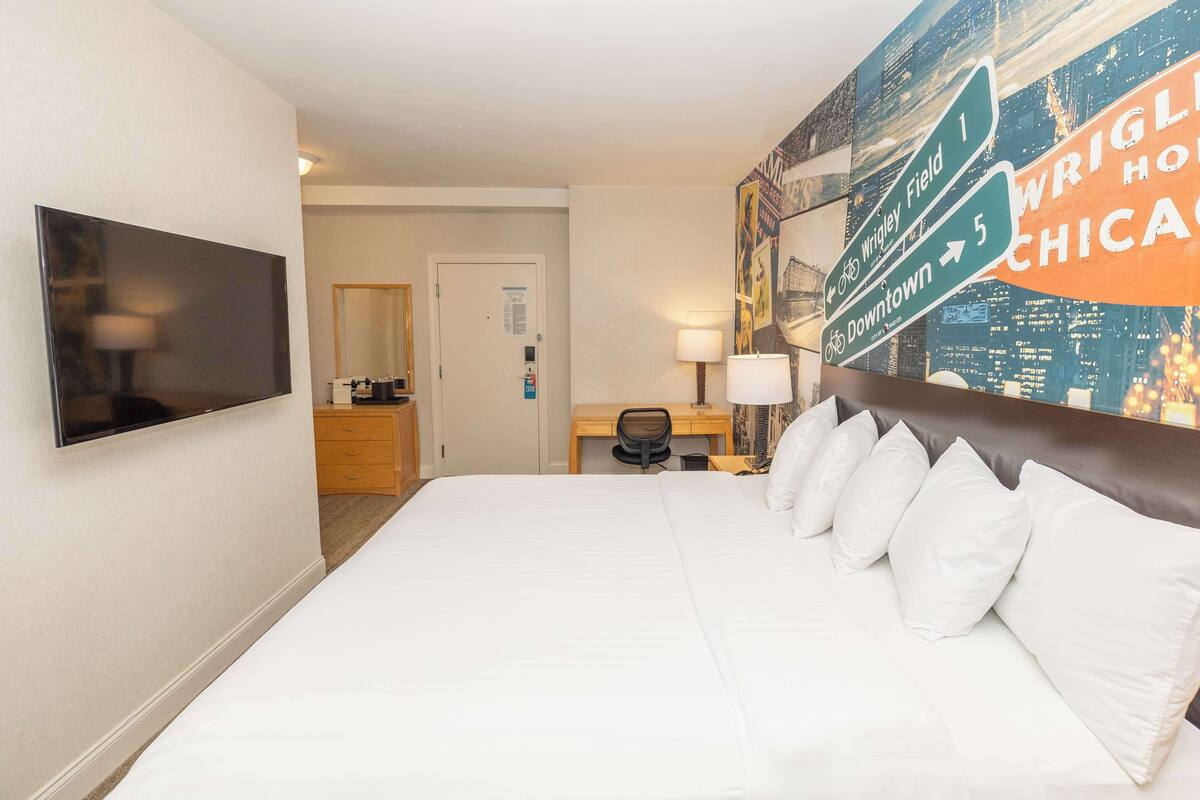 Une chambre d’hôtel avec un grand lit, un bureau, une chaise, une télévision et une fresque représentant un paysage urbain sur le mur.