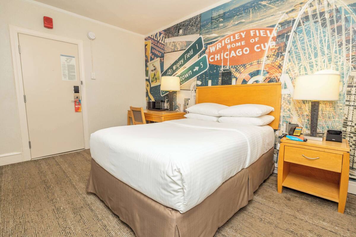 Une chambre d’hôtel avec un grand lit, deux tables de chevet avec des lampes, une fresque murale sur le thème de la ville et une porte équipée d’un détecteur d’incendie.