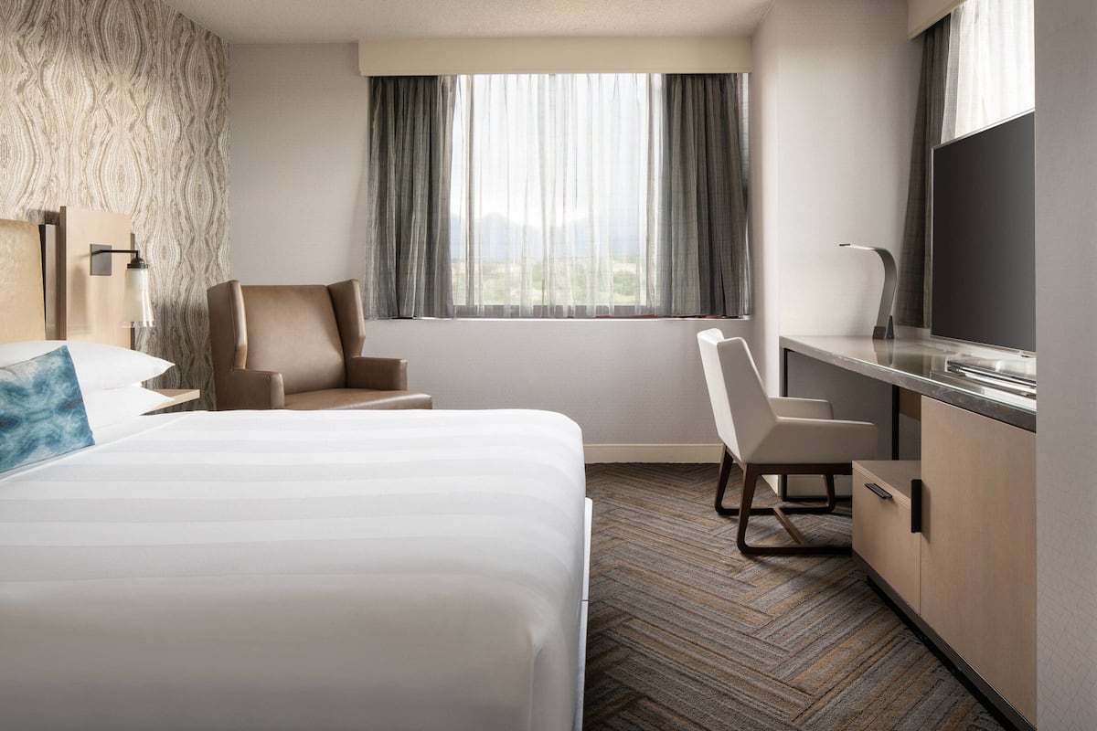 Quarto de hotel com uma cama grande, uma mesa com televisão de tela plana, uma cadeira e uma janela com cortinas.