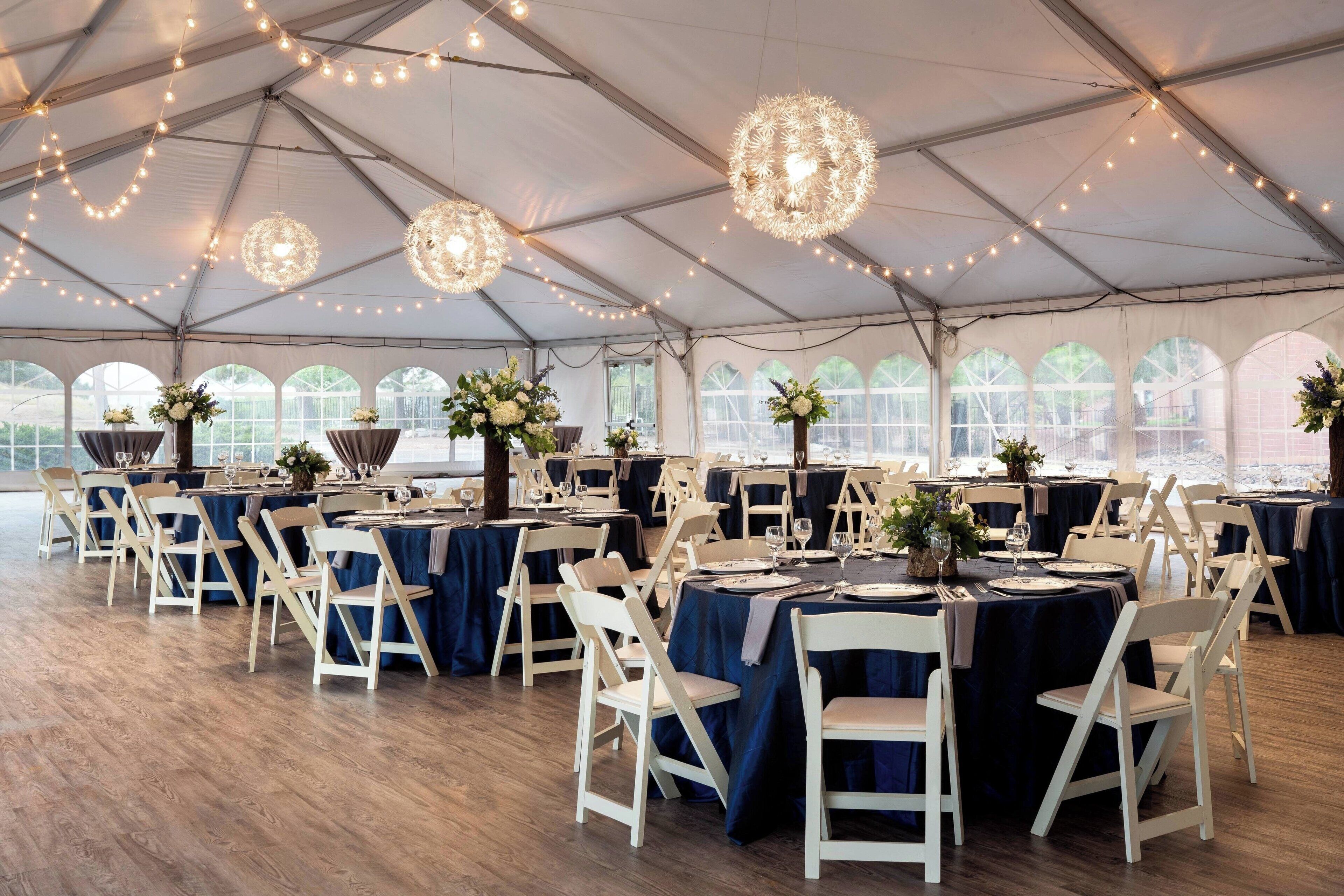 Un espacio para eventos con carpas, mesas redondas, sillas blancas y luces colgantes.