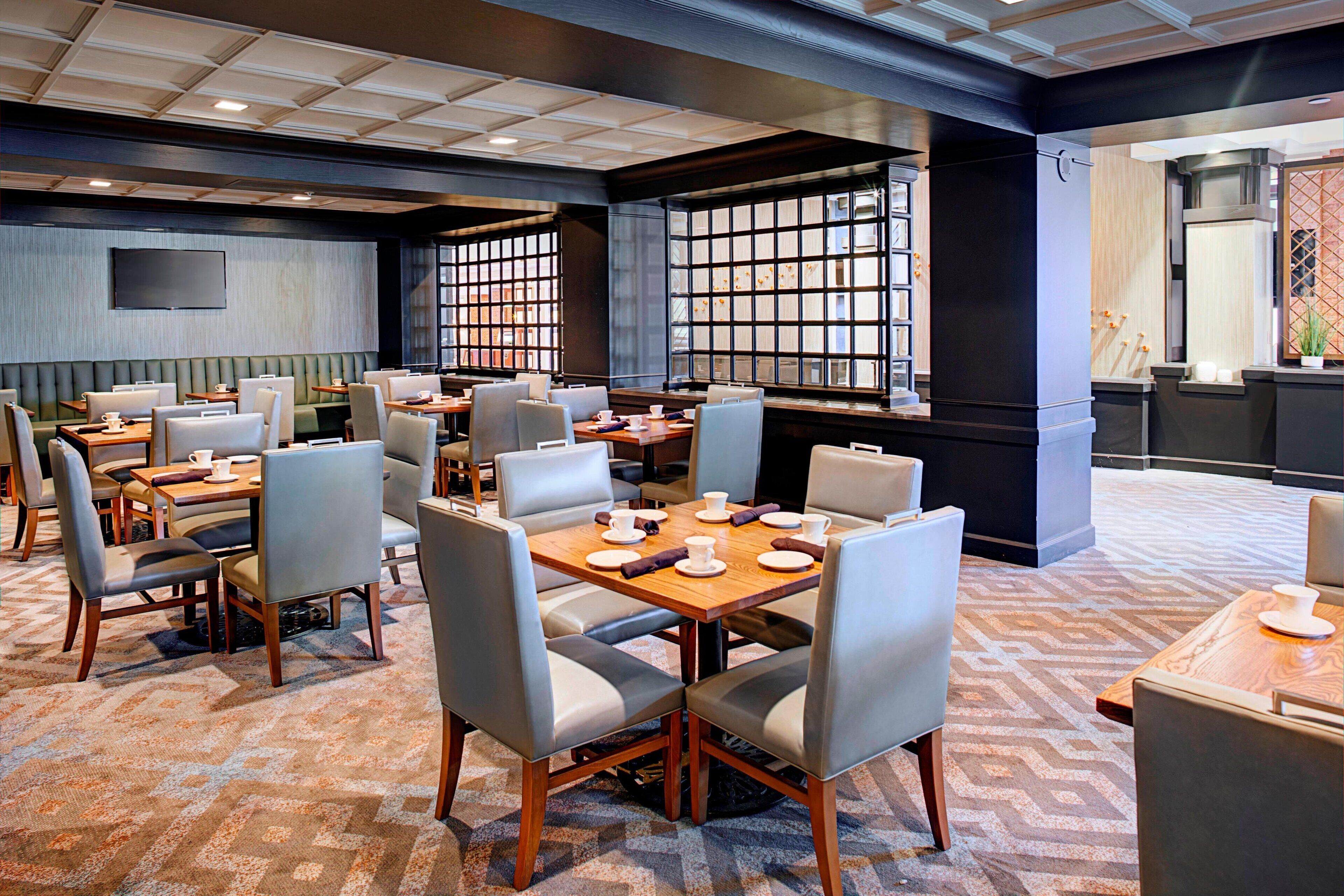Een restaurantgedeelte in een hotel met meerdere tafels en stoelen, een groot raam met uitzicht en een flatscreen aan de muur.