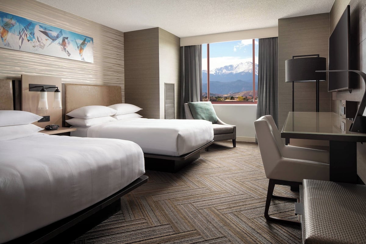 Quarto de hotel com duas camas, uma escrivaninha, uma cadeira, uma televisão e uma janela com vista para as montanhas.