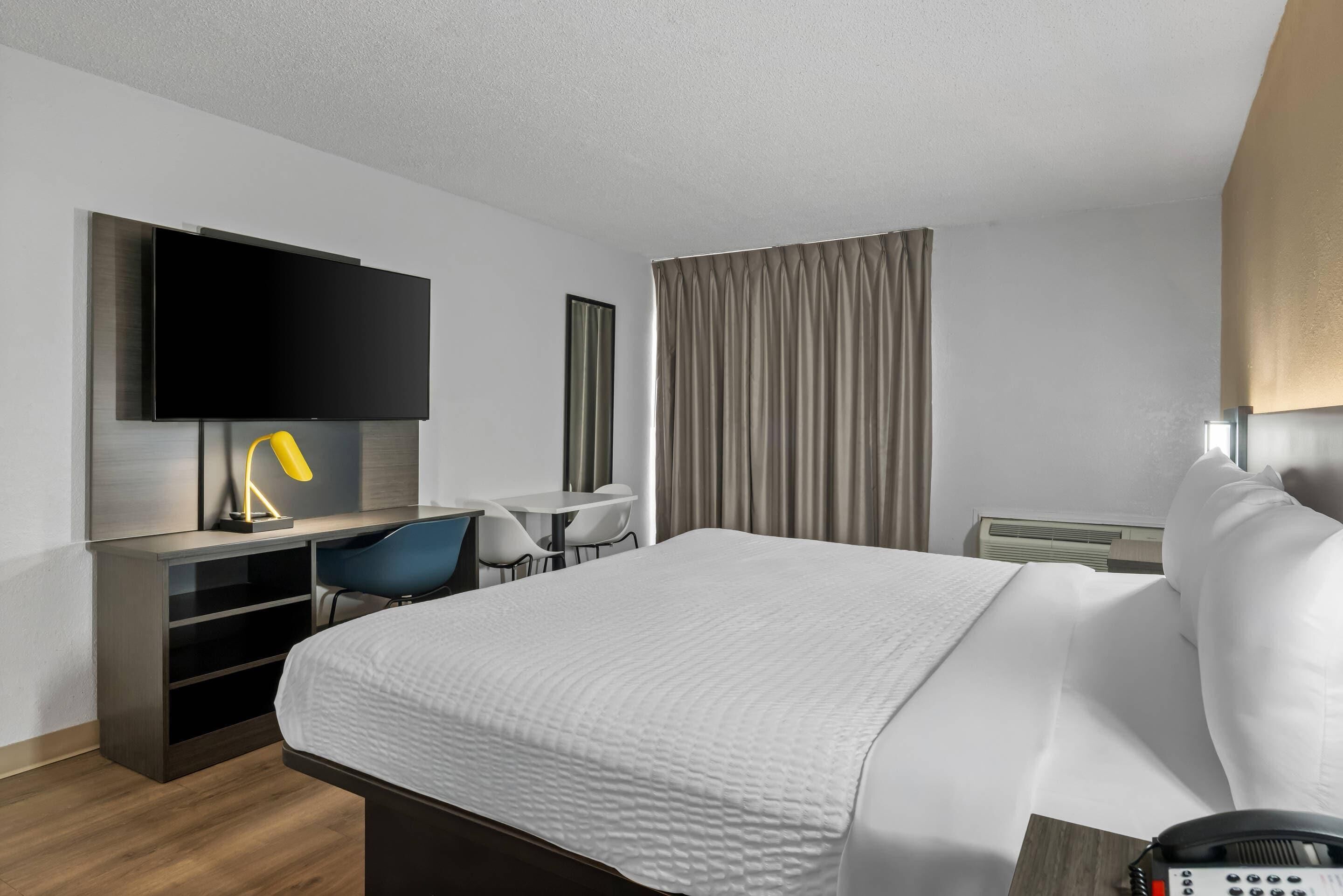 A hotel room with a bed, a desk with a chair, a flat-screen TV, a lamp, and a window with curtains.