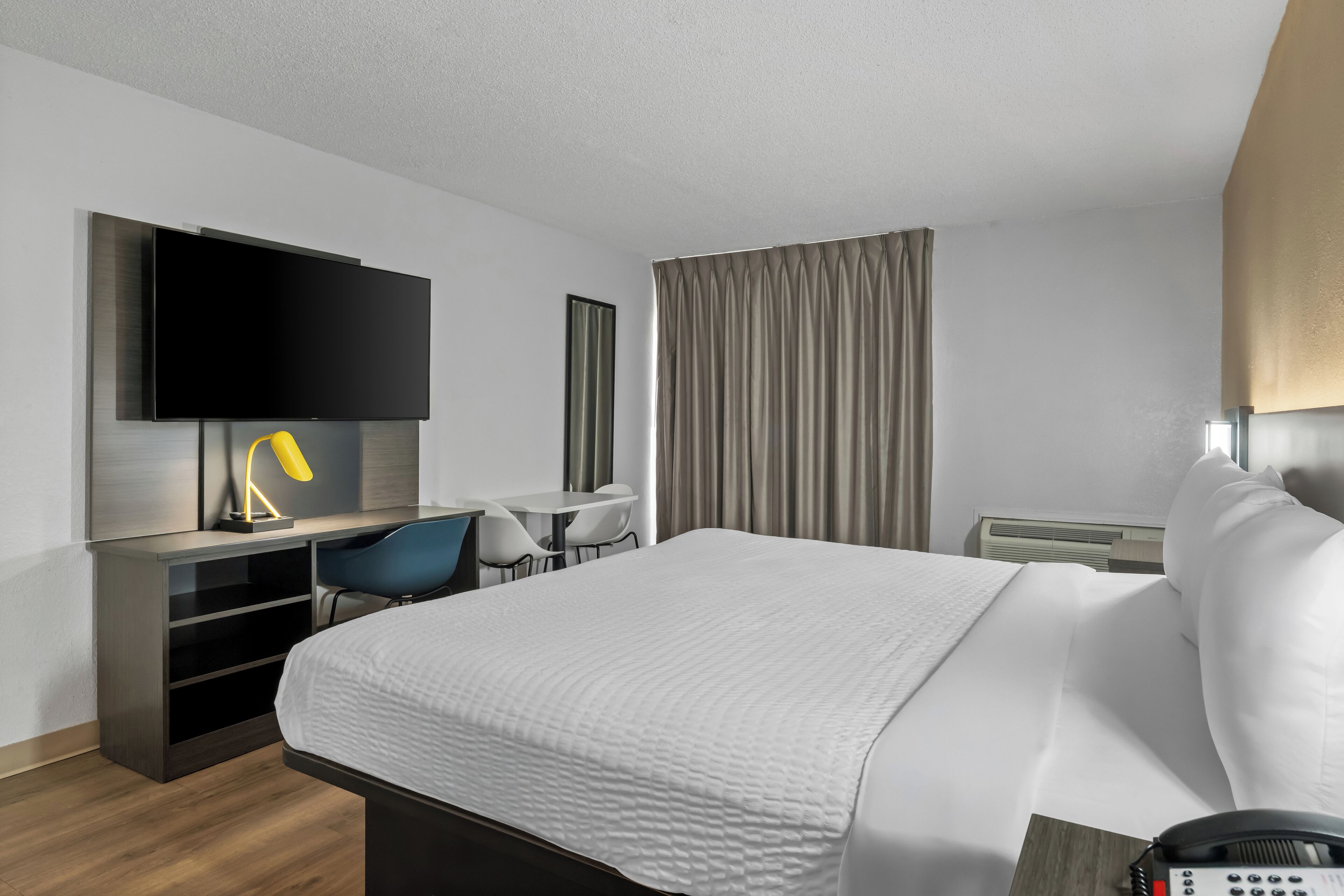 Une chambre d’hôtel moderne équipée d’un lit, d’un bureau avec une chaise, d’un téléviseur à écran plat fixé au mur et d’une grande fenêtre avec des rideaux.
