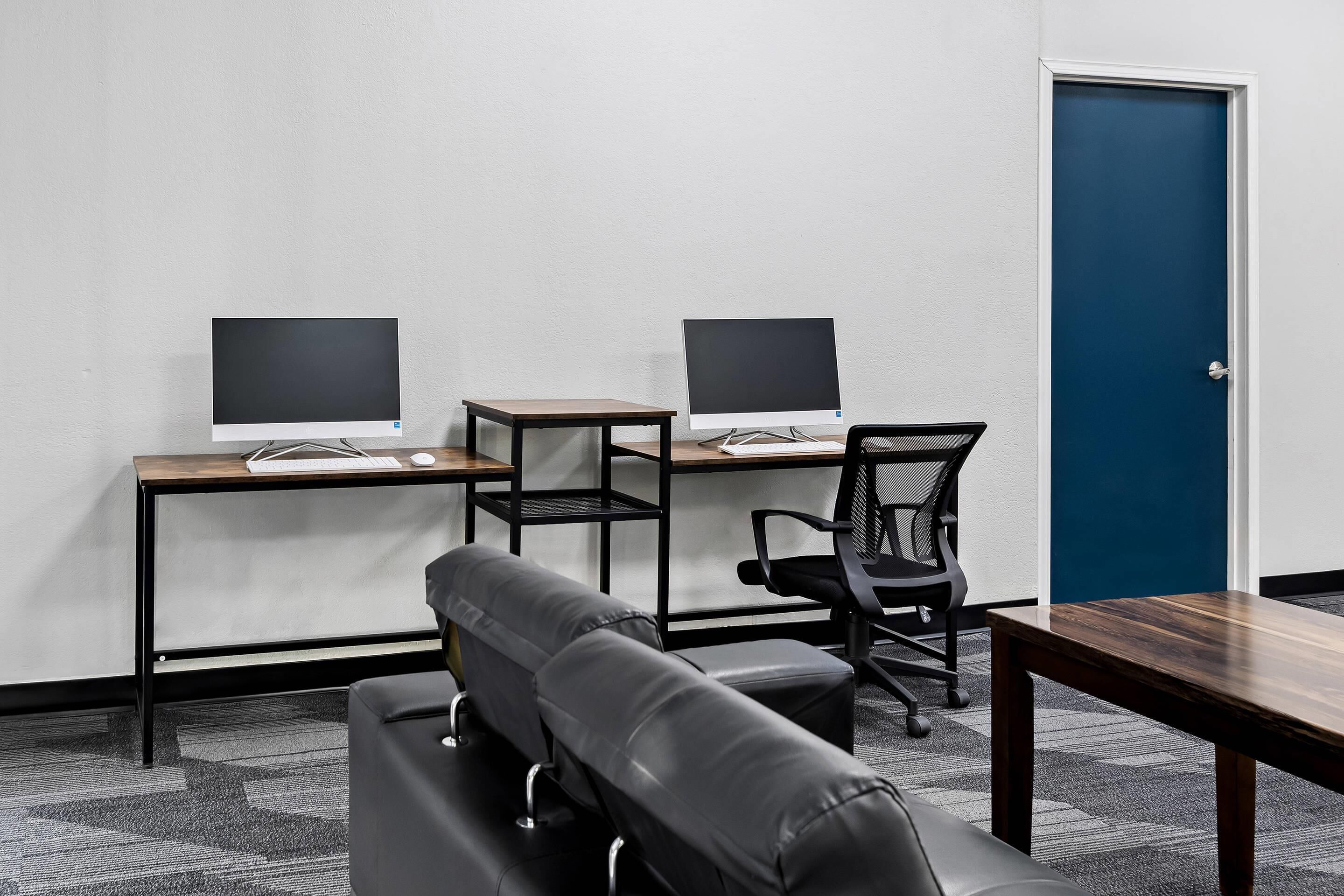 A room with two desks, each with a computer monitor, a chair, and a sofa.