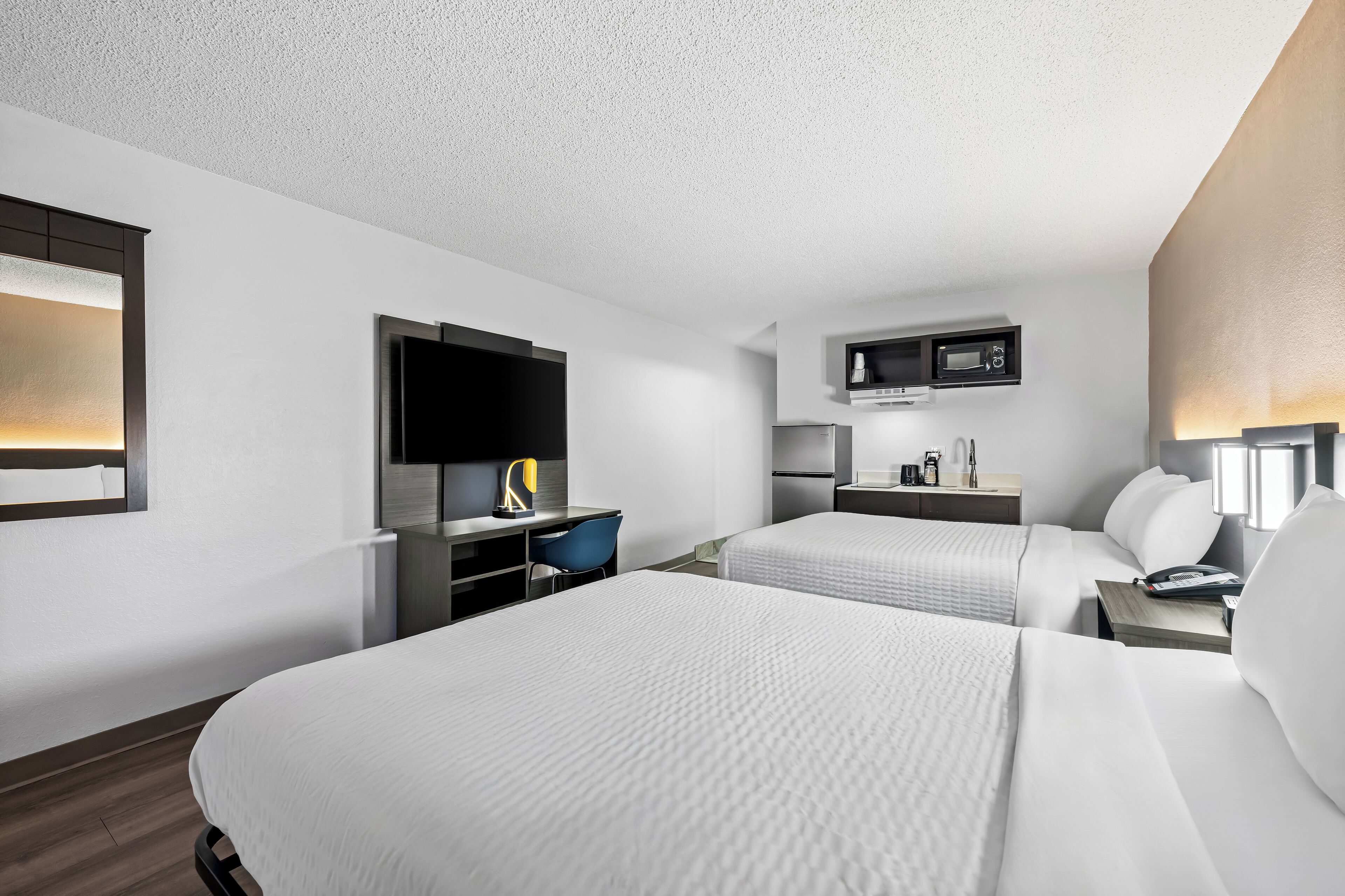 A modern hotel room with two beds, a desk, a chair, a TV, a microwave, and a small kitchenette.