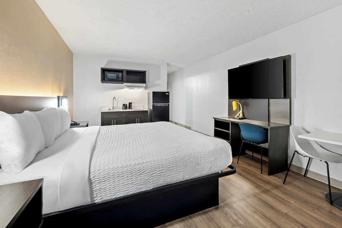 Ein modernes Hotelzimmer mit Bett, Schreibtisch, Stuhl, Küchenzeile und Fernseher.