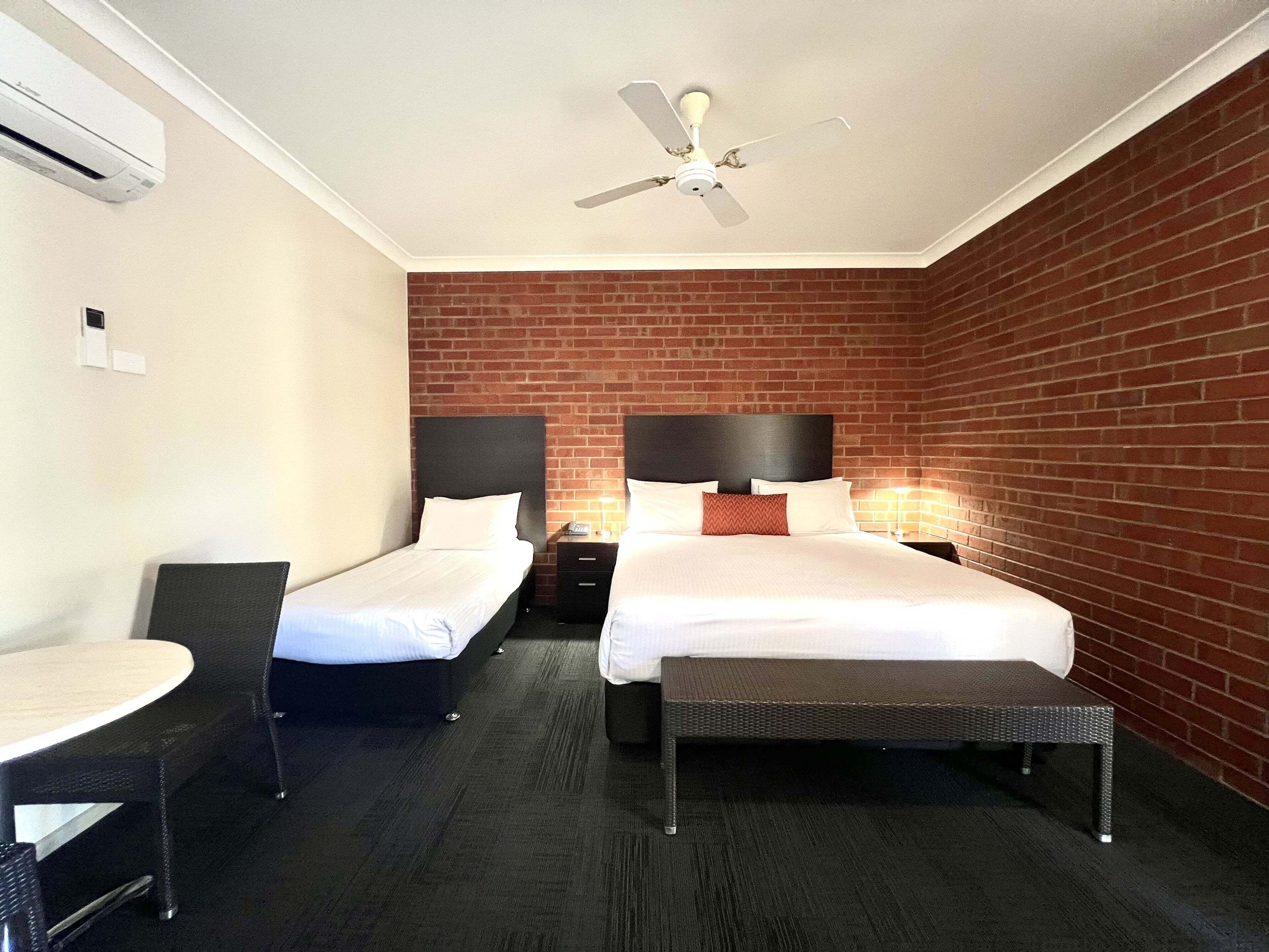 A hotel room with two beds, a desk, a chair, a ceiling fan, and a brick wall.