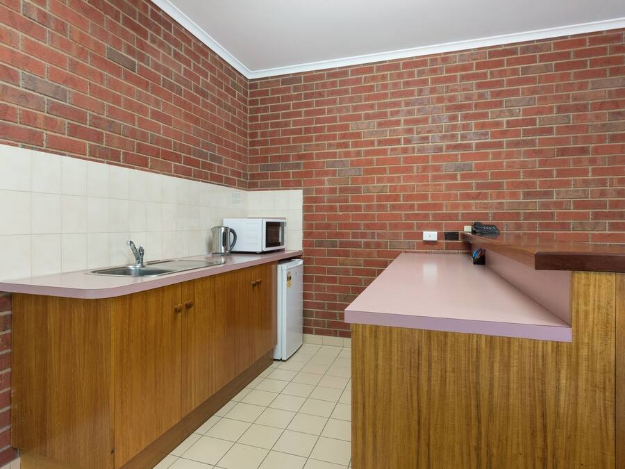 A compact kitchen with a brick wall, wooden cabinets, a white countertop, a sink, a microwave, and a refrigerator.