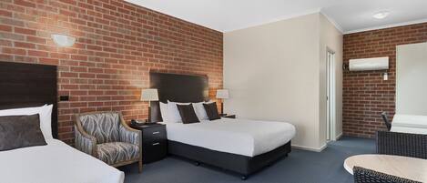 A hotel room with two beds, a brick wall, a flat-screen TV, a desk with a lamp, and a wicker chair.
