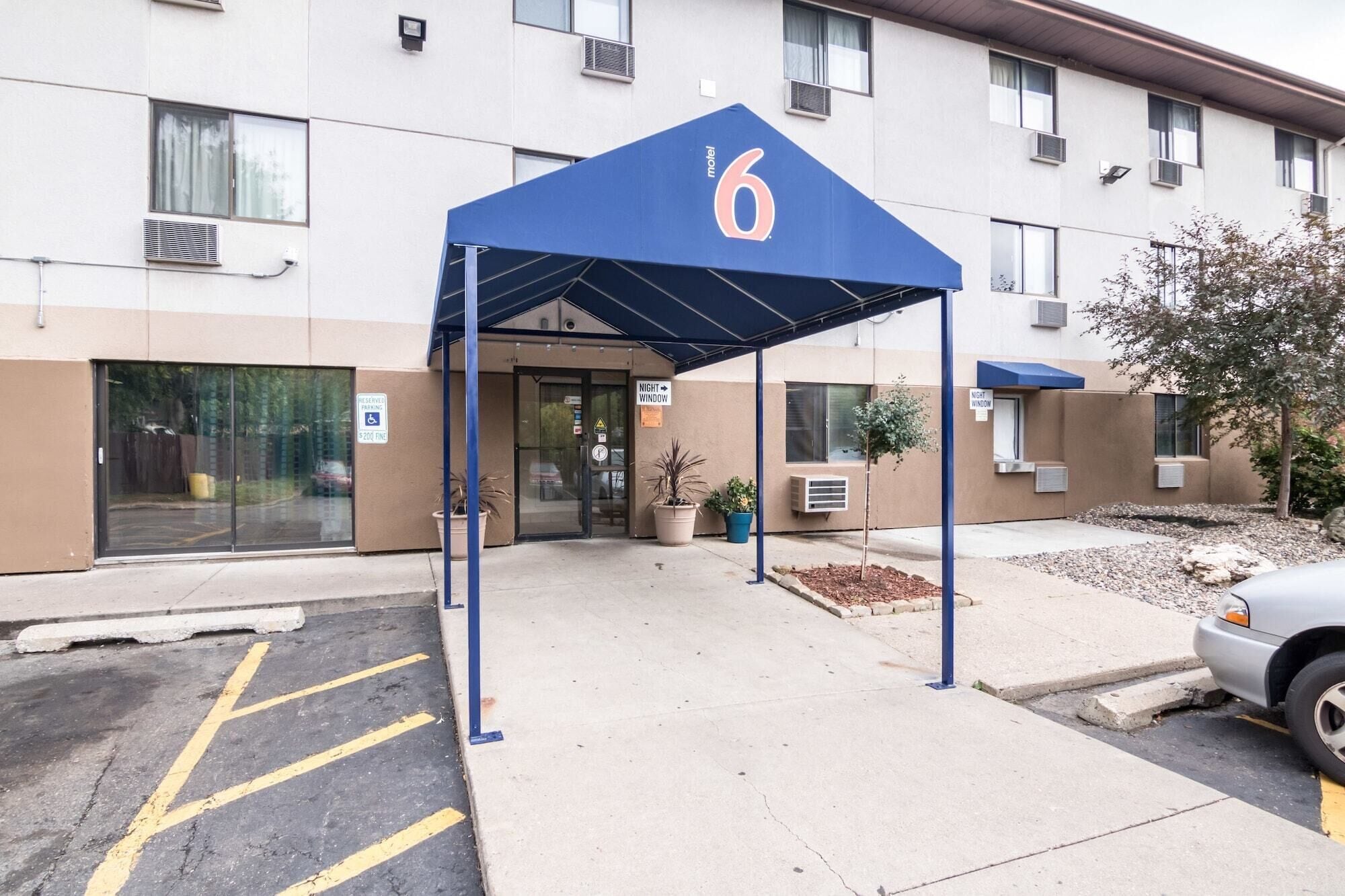 A motel with a blue awning marked with the number 6, a parking lot with marked spaces, and a building with multiple windows and air conditioning units.