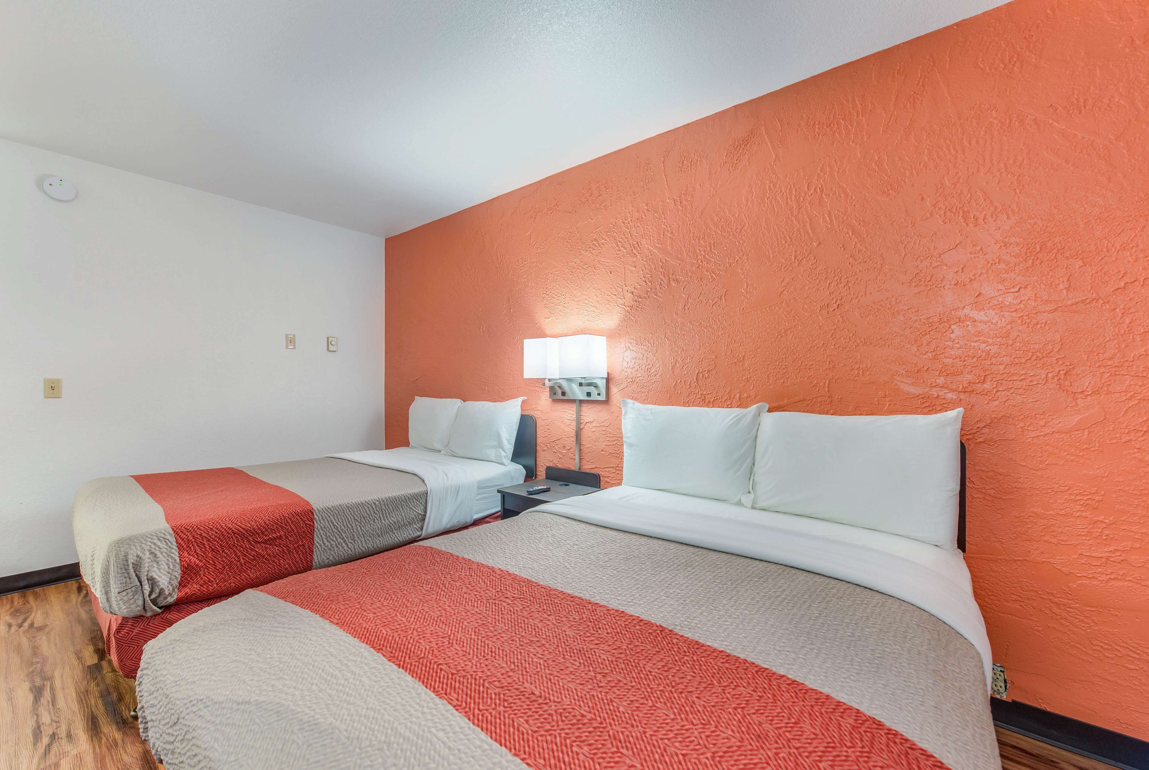 A hotel room with two beds, a nightstand, a lamp, and a wall-mounted light fixture.